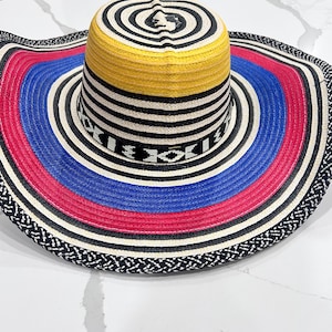 May include: A wide-brimmed straw hat featuring a vibrant color palette of yellow, blue, red, black, and white. The crown displays concentric circles, while the brim showcases alternating bands of color and a black and white woven edge pattern.