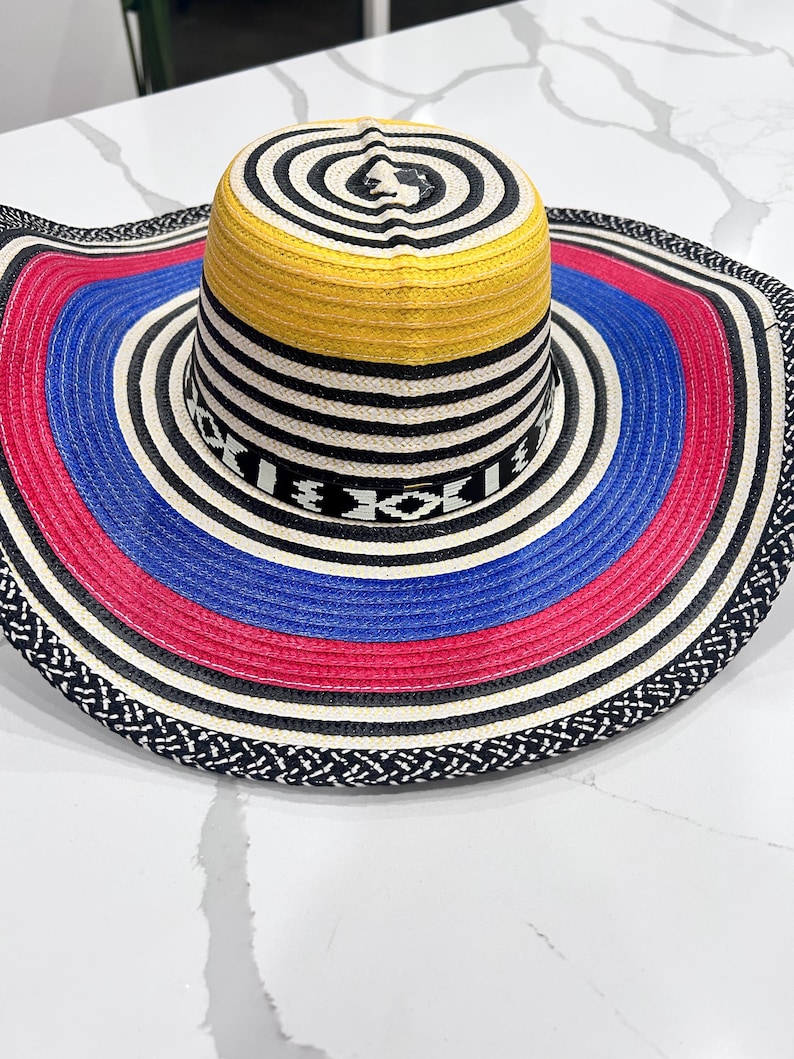 May include: A wide-brimmed straw hat featuring a vibrant color palette of yellow, blue, red, black, and white. The crown displays concentric circles, while the brim showcases alternating bands of color and a black and white woven edge pattern.