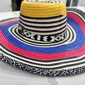 May include: A wide-brimmed hat featuring a woven texture with a yellow crown top, black and white horizontal stripes, and a decorative band with geometric patterns. The brim displays concentric rings of blue, pink, and black and white braided edges.