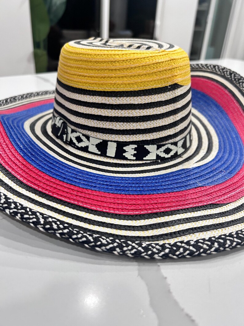 May include: A wide-brimmed hat featuring a woven texture with a yellow crown top, black and white horizontal stripes, and a decorative band with geometric patterns. The brim displays concentric rings of blue, pink, and black and white braided edges.