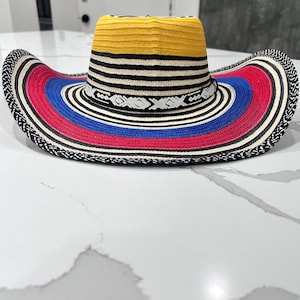 May include: A wide-brimmed hat featuring a yellow crown and a multi-colored brim with concentric rings of black, white, red, and blue. The hat includes a decorative black band with a white geometric pattern encircling the base of the crown.