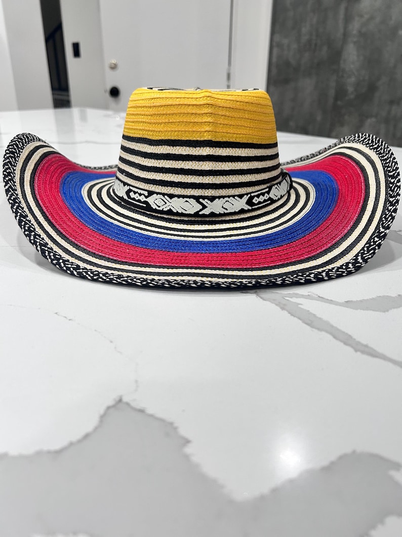 May include: A wide-brimmed hat featuring a yellow crown and a multi-colored brim with concentric rings of black, white, red, and blue. The hat includes a decorative black band with a white geometric pattern encircling the base of the crown.