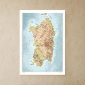 May include: A framed topographic map of Sardinia, Italy, with detailed elevation shading in shades of brown and green. The map includes place names and is set against a light blue background, with the word "Sardegna" prominently displayed.