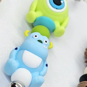 May include: Keyrings featuring cartoon monster characters. One keyring has a lime green monster with a large blue eye. Another keyring has a light blue monster with yellow horns and a white belly. Both have silver key rings.