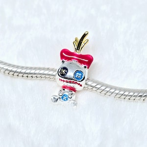 May include: A silver charm with a cartoon character design. The charm features a silver body, a red bow, and gold accents. The character has blue eyes and a stitched mouth. The charm is on a silver chain.