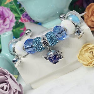 May include: A charm bracelet featuring blue and white beads, with silver accents and miniature teapot charms. The bracelet has a whimsical design, perfect for tea lovers. The beads are a mix of faceted and smooth, with a sparkling blue central bead.