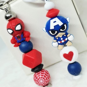 May include: Two superhero-themed keychains. One features a red Spider-Man figure, and the other has a blue Captain America figure. Each keychain has a series of colorful beads, including red, white, and blue, and a silver-colored metal ring.