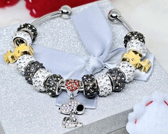 May include: Silver-toned bracelet with black, white, and clear crystal beads, gold-colored charms, and a Snoopy charm. A red heart charm is also present. The bracelet is displayed on a silver gift box with a blue ribbon.