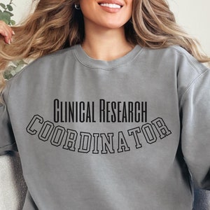 May include: A gray sweatshirt with the text "CLINICAL RESEARCH COORDINATOR" in black, arched lettering. The sweatshirt is a crew neck style and appears to be made of a soft material. The person is wearing blue jeans.