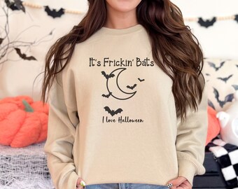May include: Beige sweatshirt with the text "It's Frickin' Bats" above a crescent moon and bats graphic. The text "I love Halloween" is below the graphic. The sweatshirt is part of a Halloween-themed collection.