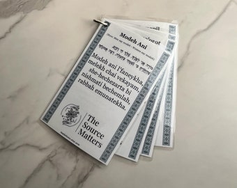 May include: A stack of white prayer cards with blue decorative borders and text. The cards are held together by a metal ring. The text includes the words "Modeh Ani" and "The Source Matters" logo. The background is a light-colored, marble-like surface.