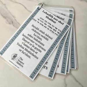 May include: A set of white cards with blue borders and Hebrew text. The cards feature a prayer for lighting Shabbat candles. The text includes the words "Lehadlik ner shel Shabbat" and "Barukh attah Adonai eloheinu melekh ha'olam".