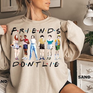 May include: A beige sweatshirt with a graphic print of the characters from the show "Stranger Things" and the text "FRIENDS DON'T LIE".