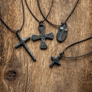 May include: Four dark gray metal pendants on thin brown cords. The pendants include a long slender cross, a wide textured cross, a smooth oval-shaped cross, and a small simple cross. Each piece features a rustic, hammered finish and dark tones.