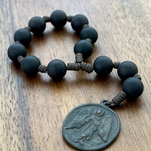 May include: A black rosary with matte finish beads and a small metal pendant. The pendant features an embossed image of an angel. The rosary is strung on a brown cord.