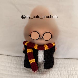 Puede incluir: Muñeco de ganchillo de un personaje con gafas redondas, una bufanda a rayas rojas y doradas y una chaqueta negra. El muñeco tiene una camisa blanca y pantalones grises. El texto "@my_cute_crochets" está en la esquina superior izquierda.