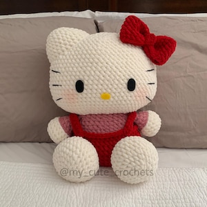 May include: White crocheted Hello Kitty plush toy with a red bow, wearing a red and pink crocheted dress. The toy is sitting on a white bedspread.