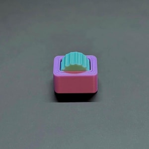 May include: A small, square object in pink and light blue. The top section has a ridged, fan-shaped design. The item is set against a grey background. The object is likely a decorative item.