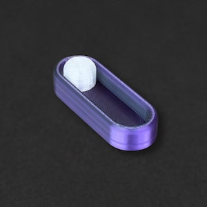 May include: A small, oval-shaped purple fidget slider with a smooth, metallic finish. The device features a recessed centre track containing a single, small, white cylindrical button that is positioned towards one end of the elongated frame.