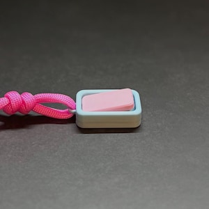May include: A small, rectangular toggle switch with a pink button and a light blue frame, attached to a pink cord. The switch is set against a dark grey background, with the cord knotted.
