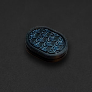 Pode incluir: Um botão de formato oval com uma superfície texturizada em azul escuro e preto, apresentando um padrão repetitivo de pequenas caveiras. O botão possui uma borda preta lisa e elevada ao redor do design central.