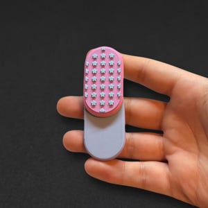 May include: A small, oval-shaped handheld brush featuring a pink top section decorated with a grid of light blue star-shaped bristles. The bottom portion of the brush is a smooth, solid light purple handle with a rounded base and flat edges.