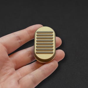 May include: An oval-shaped fidget slider with a matte gold finish and a series of six horizontal dark brown textured bars across the top surface. The object has a smooth, rounded edge and a compact design, featuring a layered construction.
