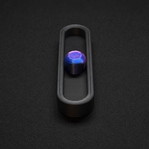 Peut inclure: Un plateau noir mat, allongé et de forme ovale, doté d'un centre évidé. À l'intérieur du plateau repose un petit objet lisse en forme de dôme, présentant un dégradé irisé vibrant aux teintes métalliques bleu, violet et magenta sur sa surface.