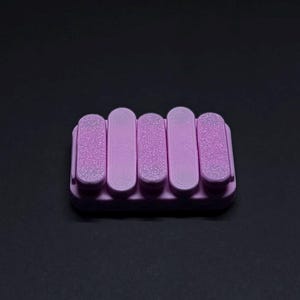 May include: A lavender soap dish with a rectangular base and five vertical, rounded bars. The bars are evenly spaced and have a textured surface. The soap dish is made of a solid material and is set against a dark background.