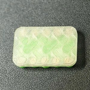 May include: A rectangular, translucent container with a green interior and a green base. The container has a series of curved, wave-like patterns inside. The object is set against a dark, textured background.