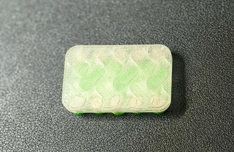 May include: A rectangular, translucent container with a green interior and a green base. The container has a series of curved, wave-like patterns inside. The object is set against a dark, textured background.