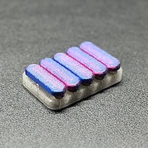May include: A set of five iridescent keycaps in shades of blue and pink, mounted on a light gray base. The keycaps have a textured surface and are arranged in a row.
