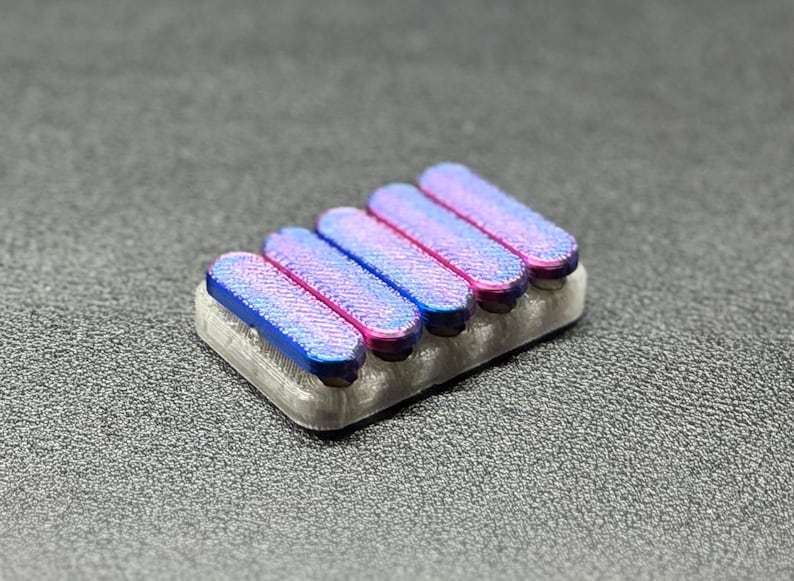 May include: A set of five iridescent keycaps in shades of blue and pink, mounted on a light gray base. The keycaps have a textured surface and are arranged in a row.