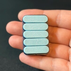 May include: A rectangular, handheld device with five light blue, textured, oval-shaped buttons. The device has a dark gray frame and is held in a person's hand. The buttons are evenly spaced and slightly raised.