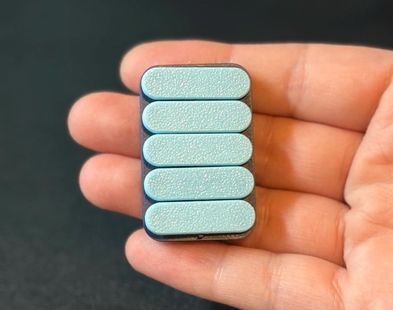 May include: A rectangular, handheld device with five light blue, textured, oval-shaped buttons. The device has a dark gray frame and is held in a person's hand. The buttons are evenly spaced and slightly raised.
