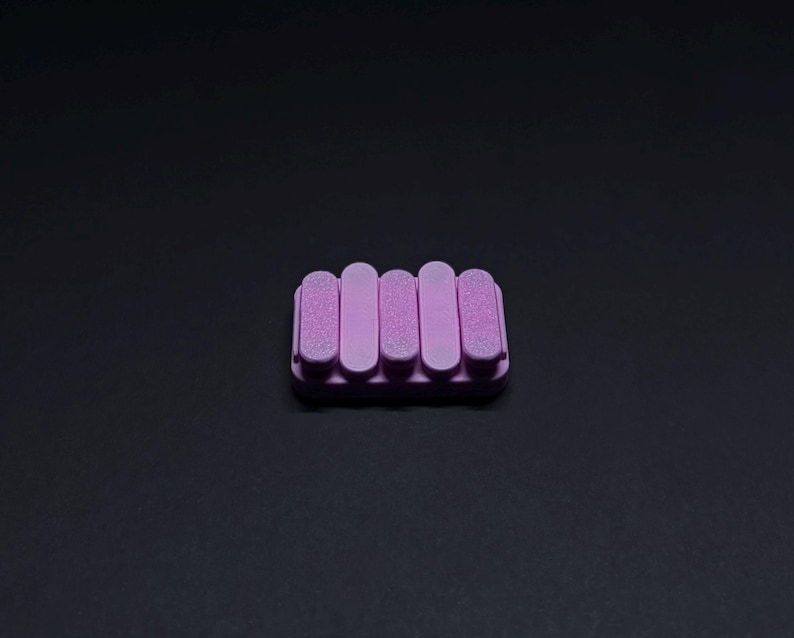 May include: A lavender soap dish with a rectangular base and five vertical, rounded bars. The bars are evenly spaced and have a textured surface. The soap dish is made of a solid material and is set against a dark background.