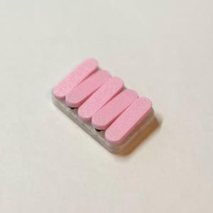 May include: A small, rectangular nail file with five pink, rectangular abrasive pads. The pads are arranged in a row and are attached to a light gray base. The nail file is designed for manicure and pedicure use.