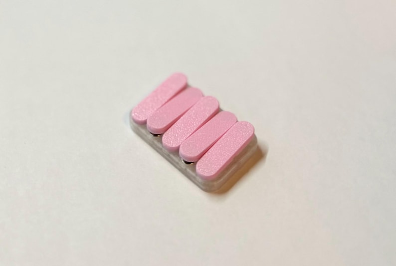 May include: A small, rectangular nail file with five pink, rectangular abrasive pads. The pads are arranged in a row and are attached to a light gray base. The nail file is designed for manicure and pedicure use.