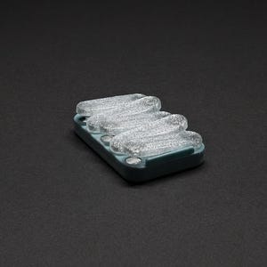 May include: A rectangular, teal-colored soap dish with three clear, wavy soap bars. The soap bars have a glittery, textured appearance. The dish is set against a dark gray background.