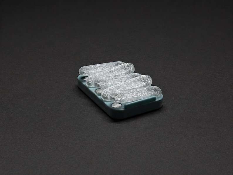 May include: A rectangular, teal-colored soap dish with three clear, wavy soap bars. The soap bars have a glittery, textured appearance. The dish is set against a dark gray background.
