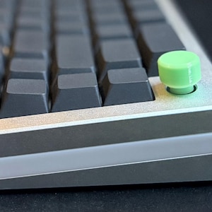 May include: A close-up of a mechanical keyboard with dark grey keycaps and a light green accent key. The keyboard has a silver frame with a light grey accent. The keycap is a bright, solid green colour.