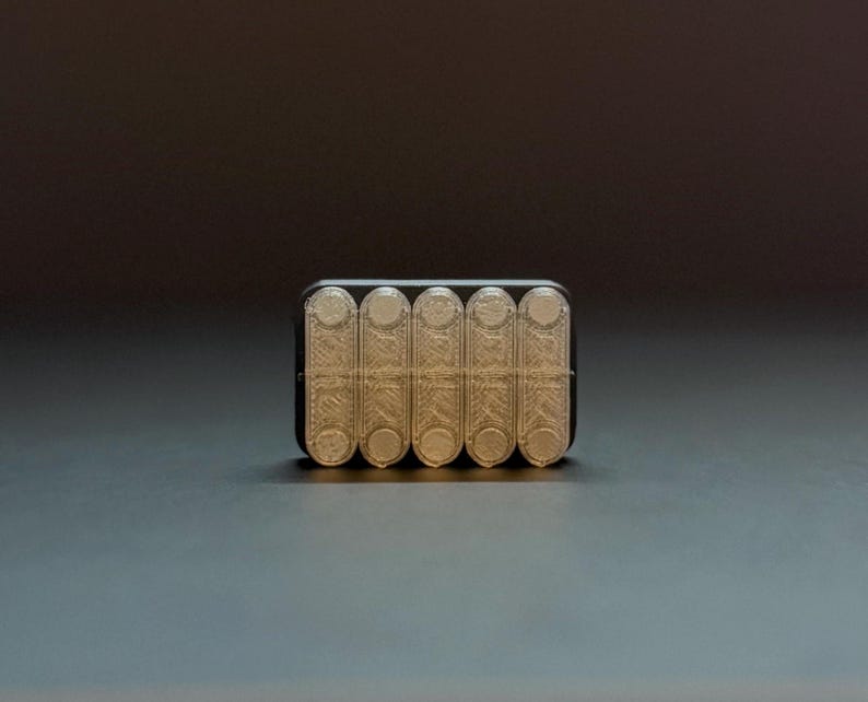 May include: A rectangular, black and beige object with six cylindrical compartments. The compartments are beige and have a textured, patterned design. The object is on a dark gray surface.