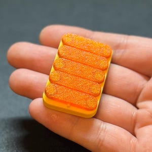 May include: A small, rectangular, yellow object with six orange, textured, oblong shapes arranged in parallel. The object is held in a person's hand, with a dark background.