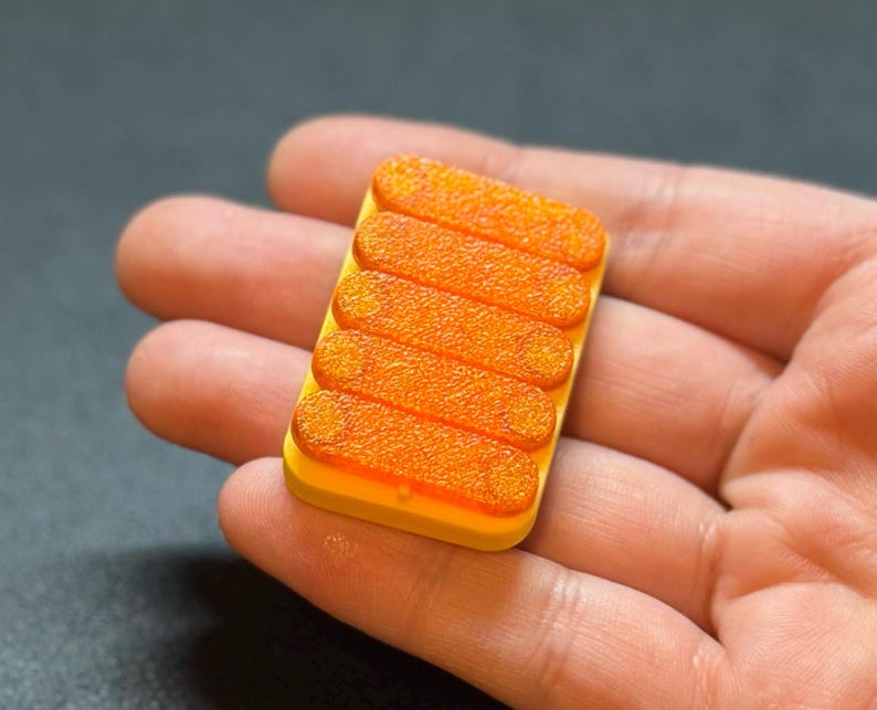 May include: A small, rectangular, yellow object with six orange, textured, oblong shapes arranged in parallel. The object is held in a person's hand, with a dark background.