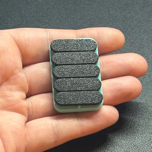 May include: A small, rectangular, mint-green object with six black, textured, oval pads. The pads are arranged in a vertical row. The object is held in a person's hand.