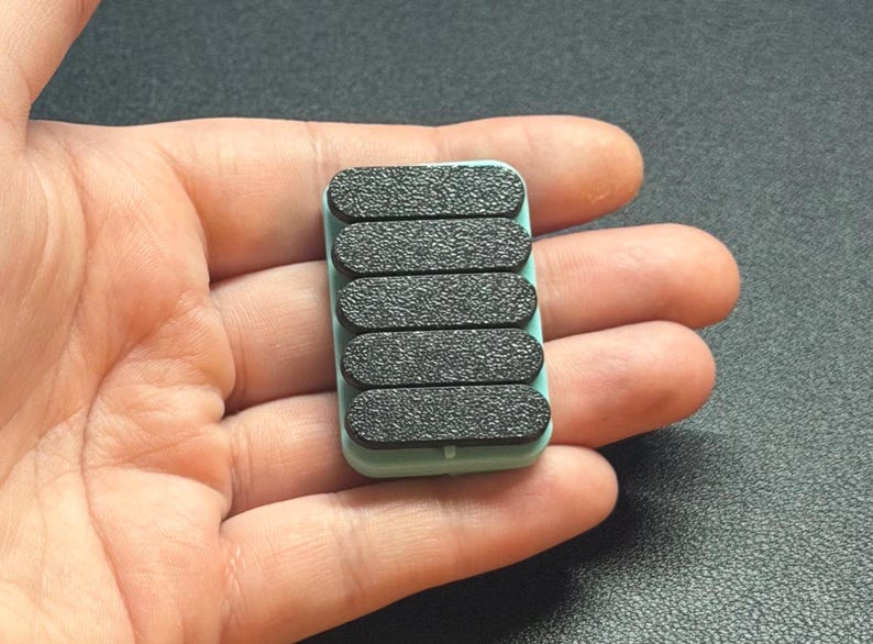 May include: A small, rectangular, mint-green object with six black, textured, oval pads. The pads are arranged in a vertical row. The object is held in a person's hand.