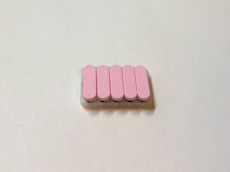 May include: A pink nail file with six rectangular pink pads. The pads are arranged in a row on a clear base. The nail file is designed for smoothing and shaping nails. The background is plain white.