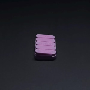May include: A stack of six lavender-colored nail files in a rectangular case. The files are arranged in a neat, stacked formation, with a slightly textured surface. The case is also lavender, and the background is a solid black.