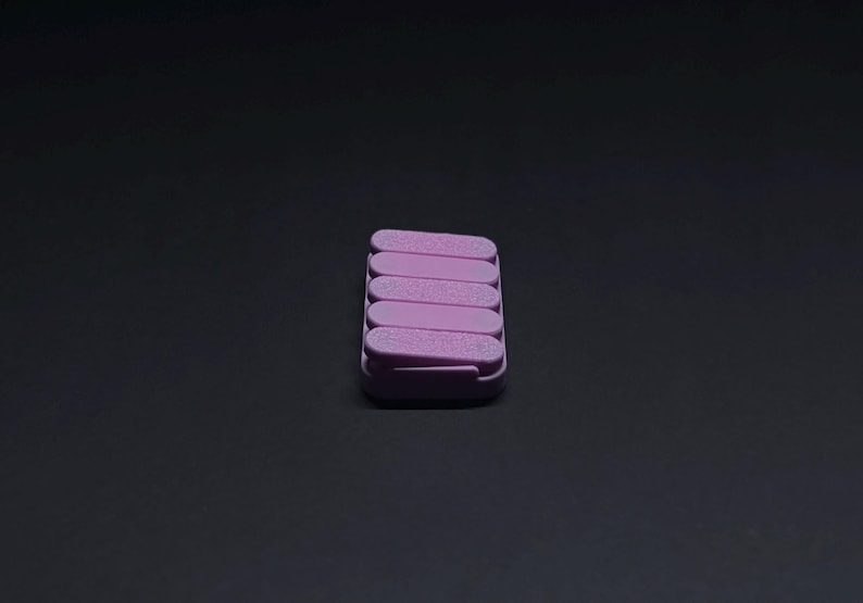 May include: A stack of six lavender-colored nail files in a rectangular case. The files are arranged in a neat, stacked formation, with a slightly textured surface. The case is also lavender, and the background is a solid black.
