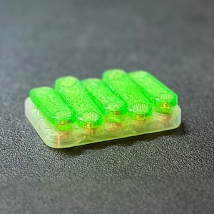 May include: A small, rectangular object with five bright green, cylindrical components arranged in a row. The base is a translucent, light green color. The object is set against a dark gray background.